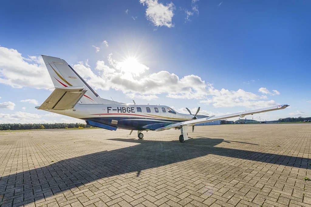A white and blue TBM aircraft with registration F-HBGE parked on a patterned paved surface under a bright sunny sky.