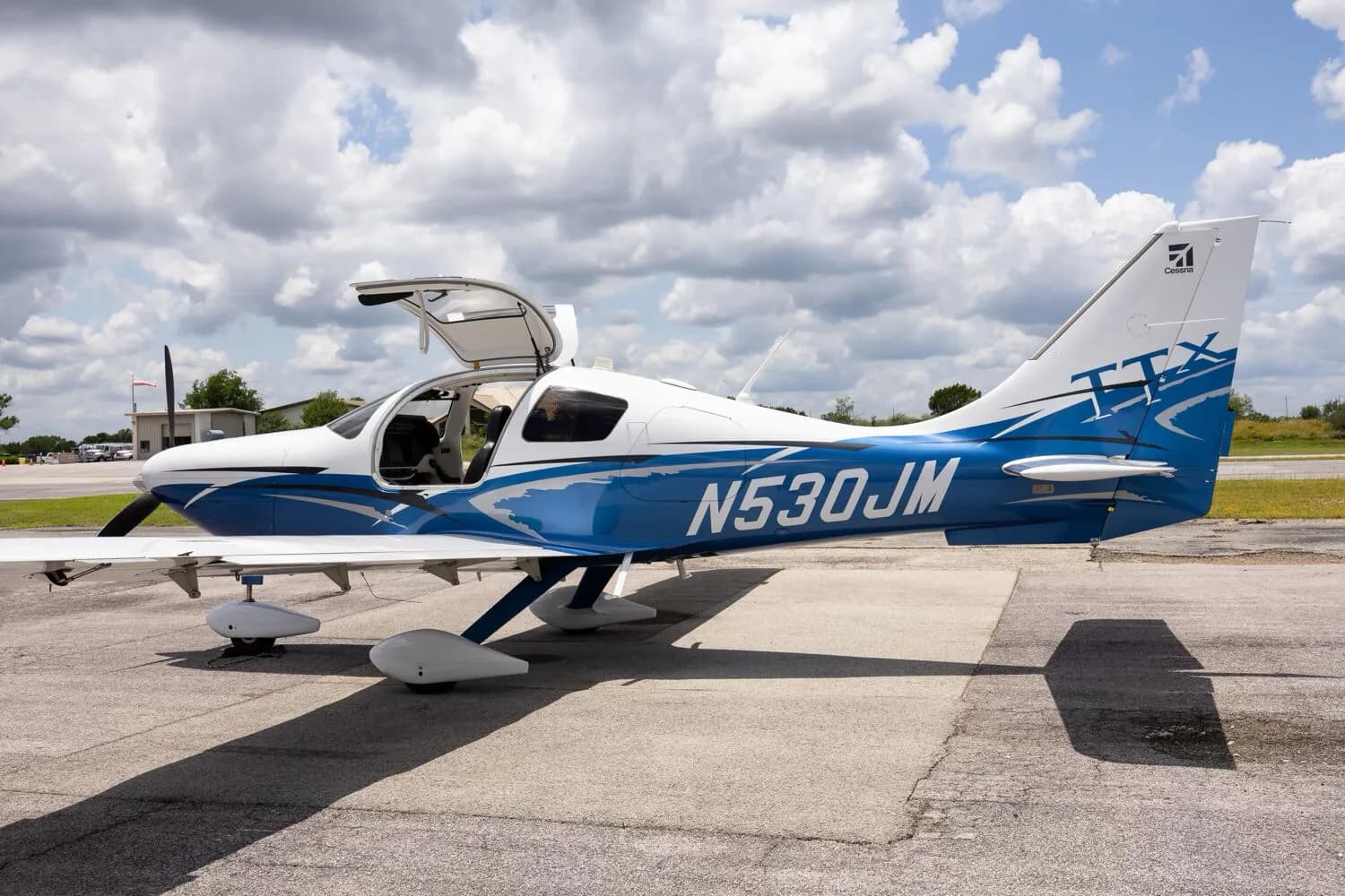 A side view of the white and blue Cessna TTX N530JM with its gull-wing doors open, parked on a tarmac.