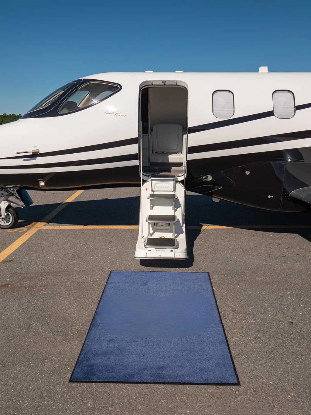 A close-up of the front fuselage of a white and black HondaJet with its main entry door open and a blue mat on the tarmac.