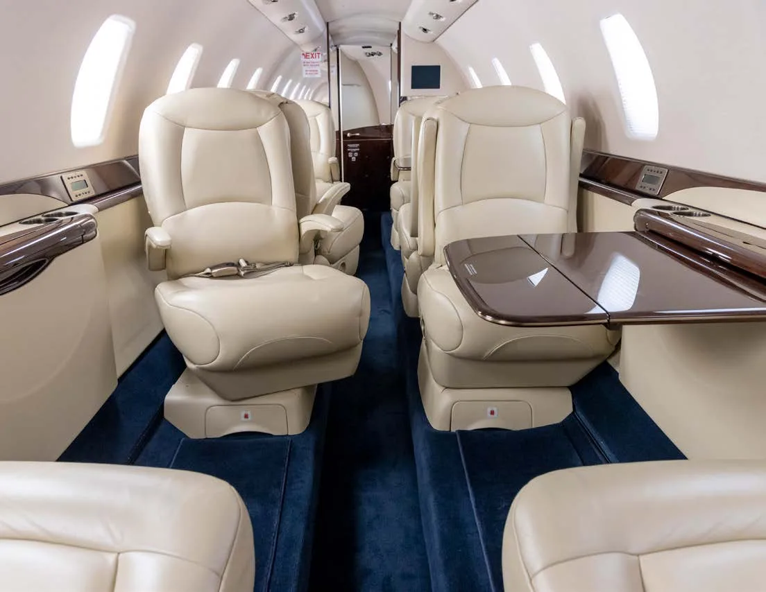 The luxurious interior cabin of a business jet with cream leather seats, dark wood accents, and blue carpet.