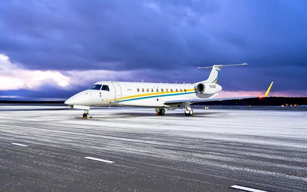 A white Embraer Legacy 600 business jet with blue and yellow stripes on a snowy tarmac under a dramatic sky.