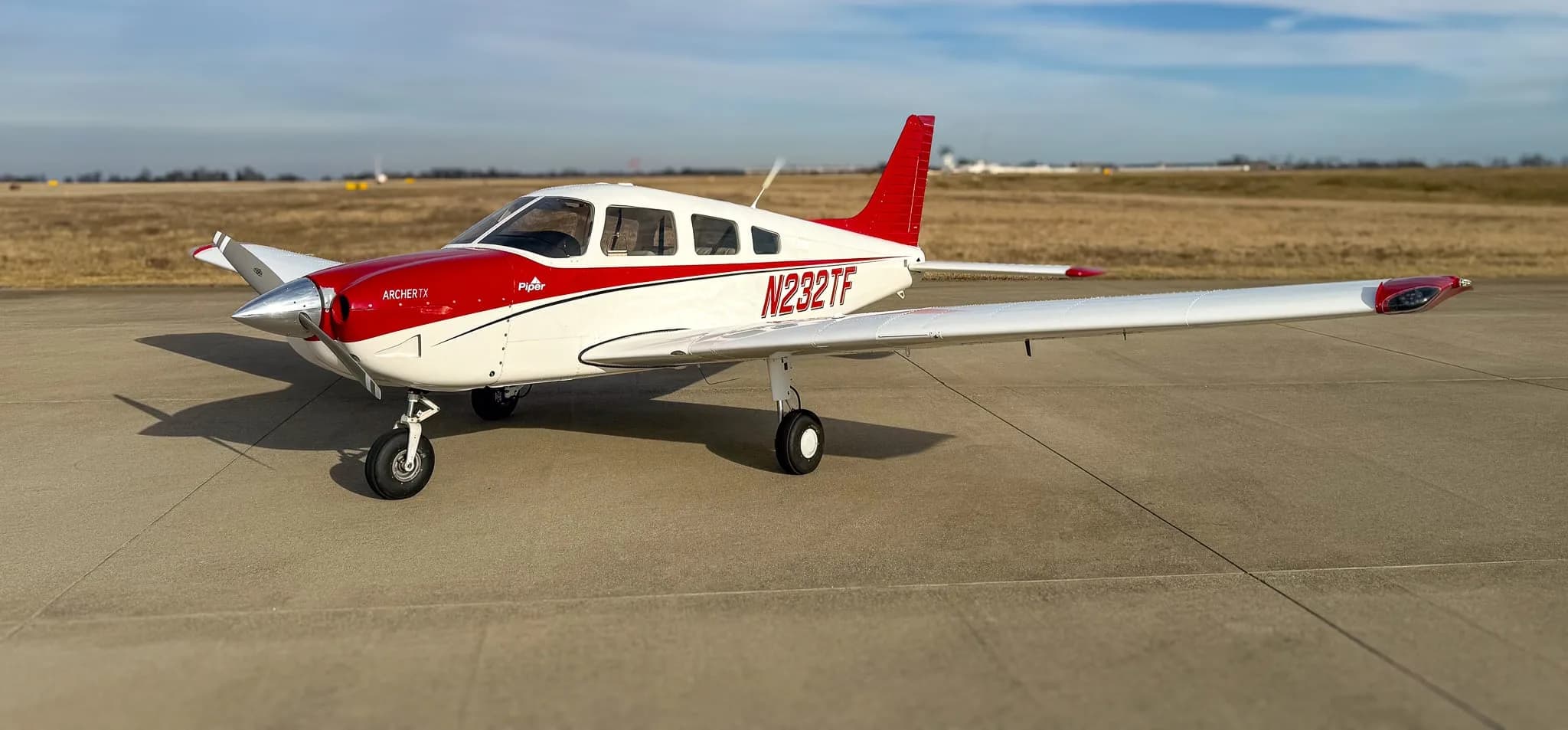 A white and red Piper Archer TX aircraft, tail number N232TF, parked on an airfield under a partly cloudy sky.