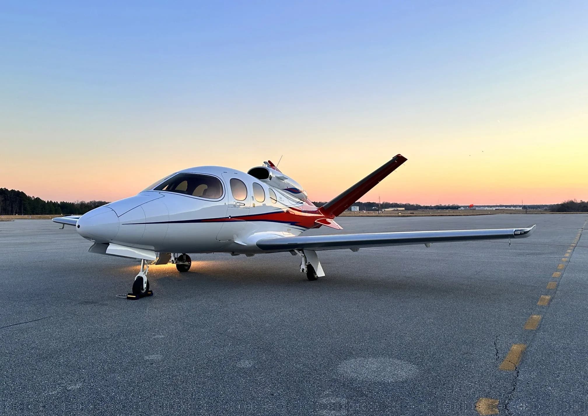 A white Cirrus Vision Jet with red and black accents is parked on an asphalt tarmac at sunset, with its landing lights illuminated.