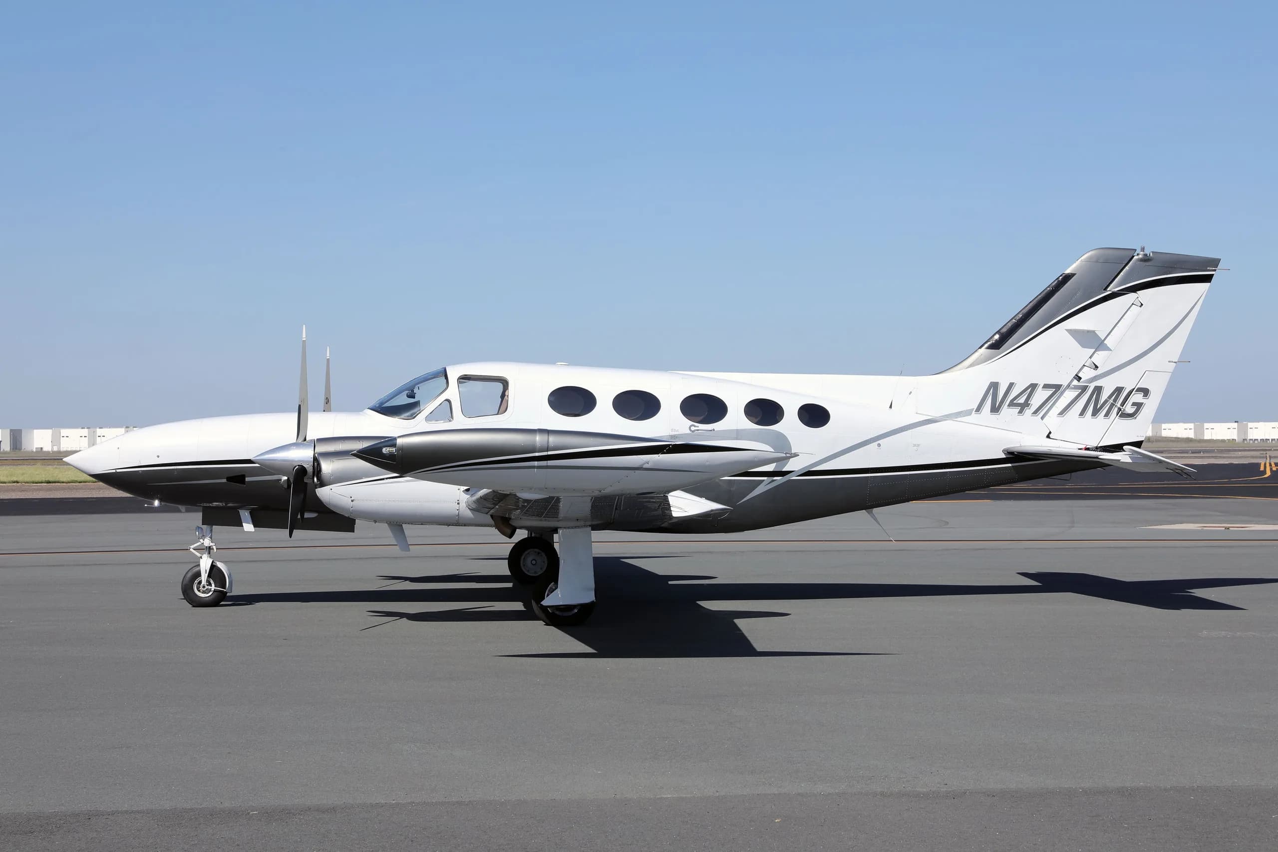 A white and grey twin-engine Cessna 414 Chancellor aircraft, N477MG, parked on an asphalt tarmac under a clear sky.