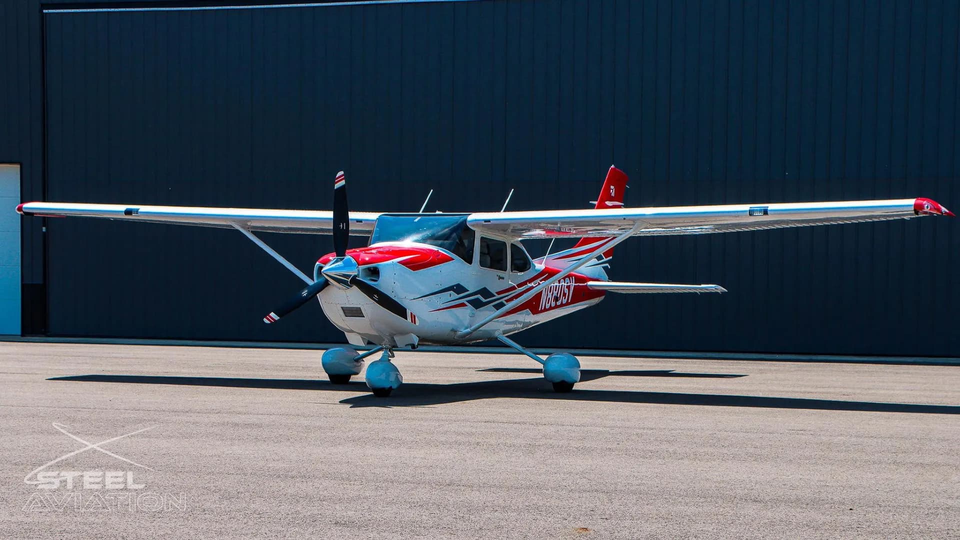 A three-quarter front view of a white Cessna T182T Skylane TC with red and grey stripes, parked on an asphalt tarmac in front of a dark hangar.