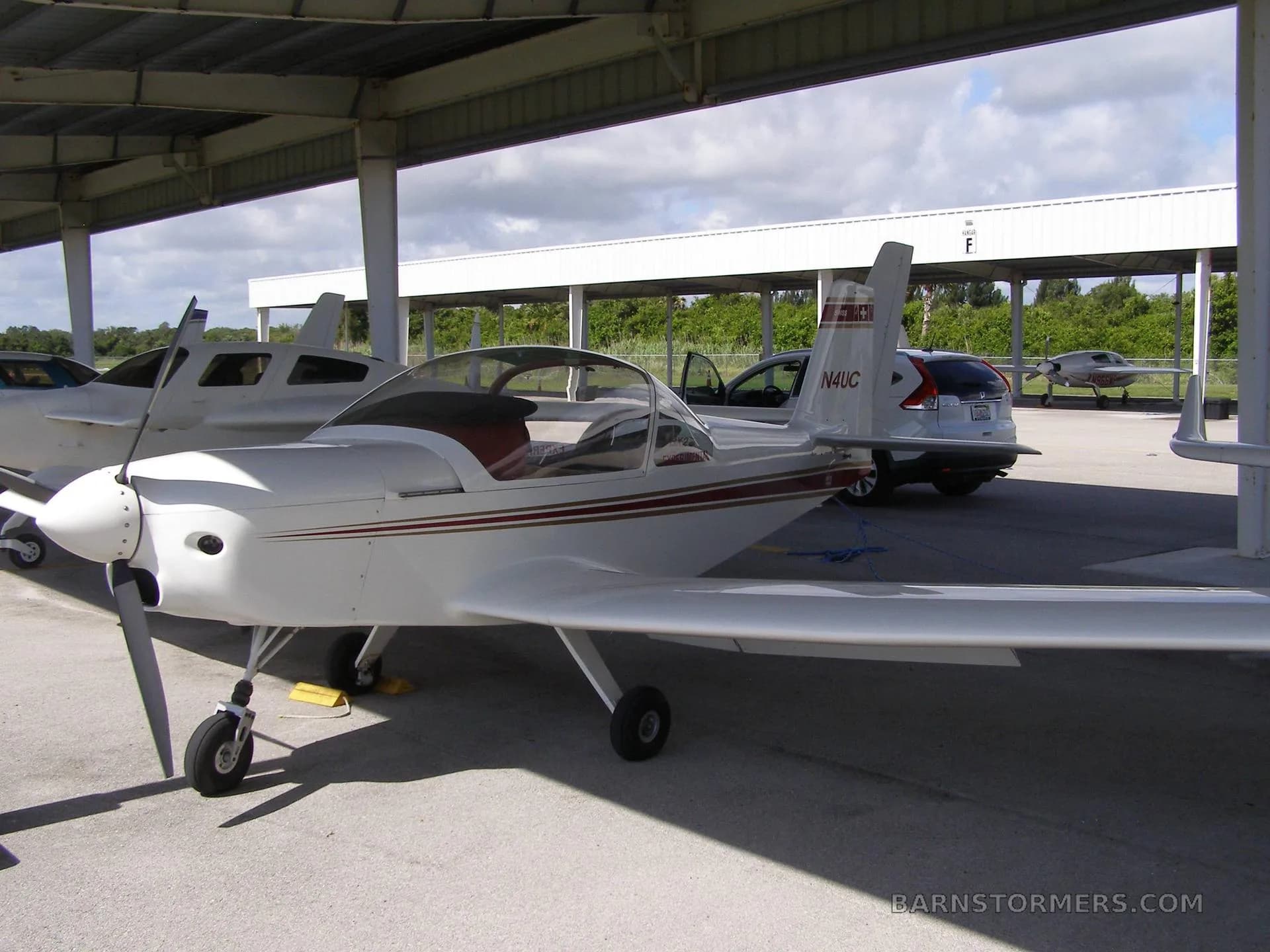 A white experimental aircraft with red and gold stripes, registration N4UC, parked under a hangar with a car and other aircraft nearby.