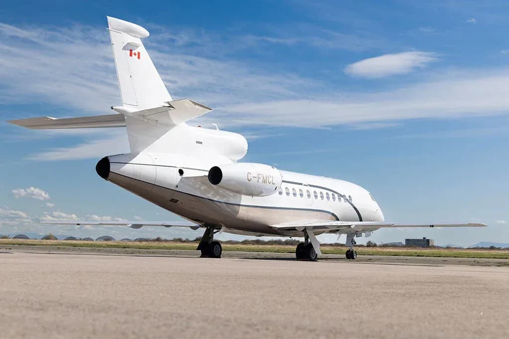 A white Dassault Falcon business jet with a brown stripe, registration C-FMCL, viewed from the rear on an airfield.