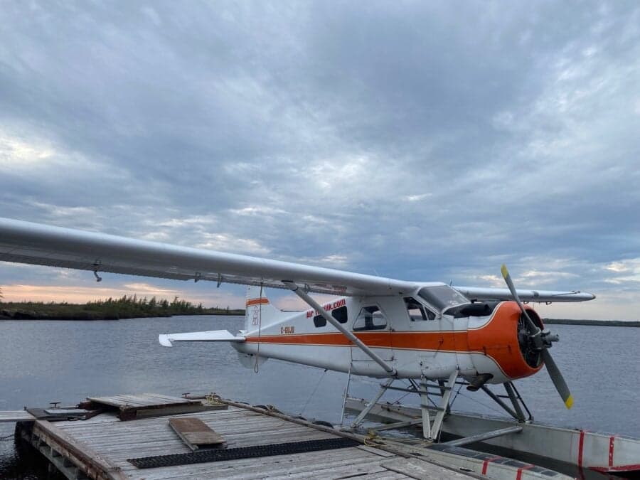 A de Havilland Beaver floatplane is docked on a wooden platform on a lake under a cloudy sky.