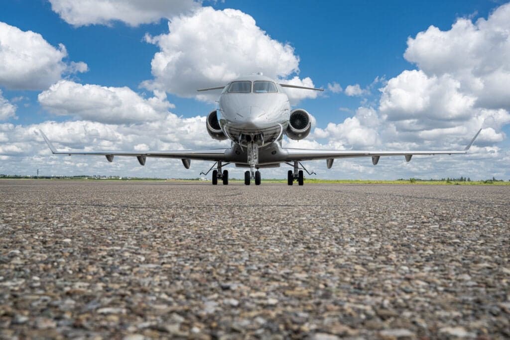 A silver and gray Challenger aircraft sits on the tarmac under a blue sky with clouds, facing forward.
