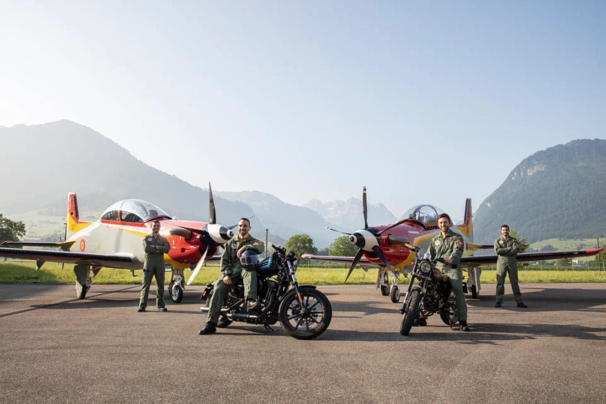Two Pilatus PC-7 MKX aircraft with people and motorcycles on a runway.
