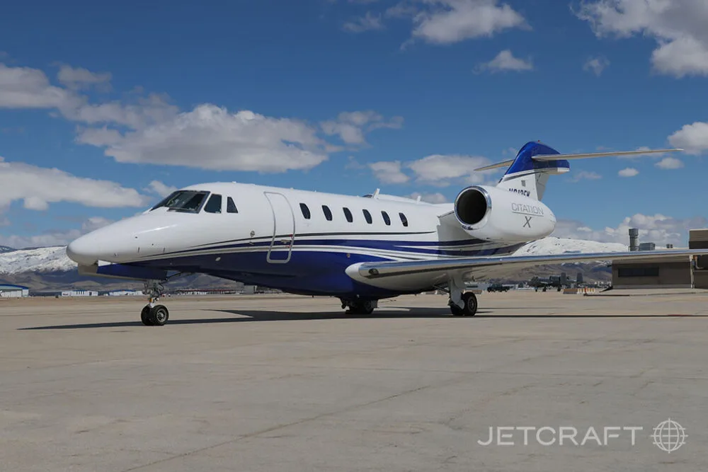 A white and blue Cessna Citation X business jet is parked on an airfield under a clear blue sky.