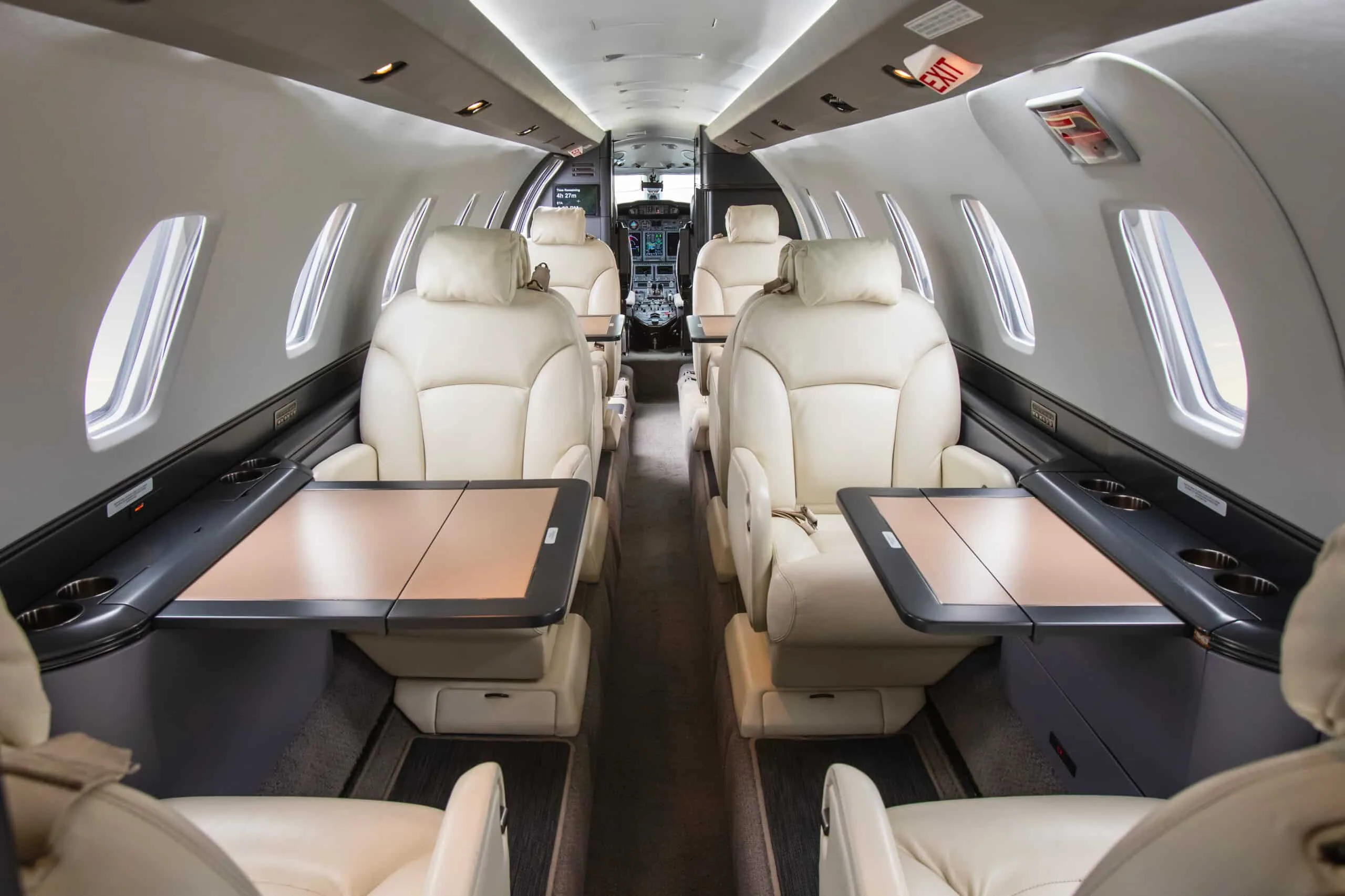 A view down the aisle of a private jet cabin, showing light-colored leather seats, fold-out tables, and a glimpse of the cockpit.
