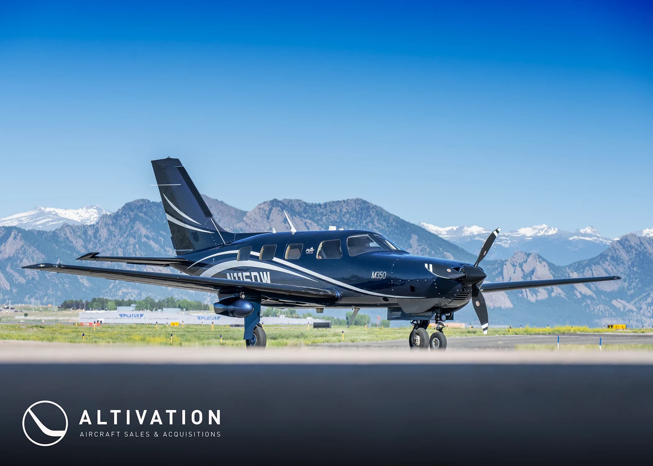 A dark blue Piper M350 aircraft with white and grey accents parked on an airfield with snow-capped mountains in the background.