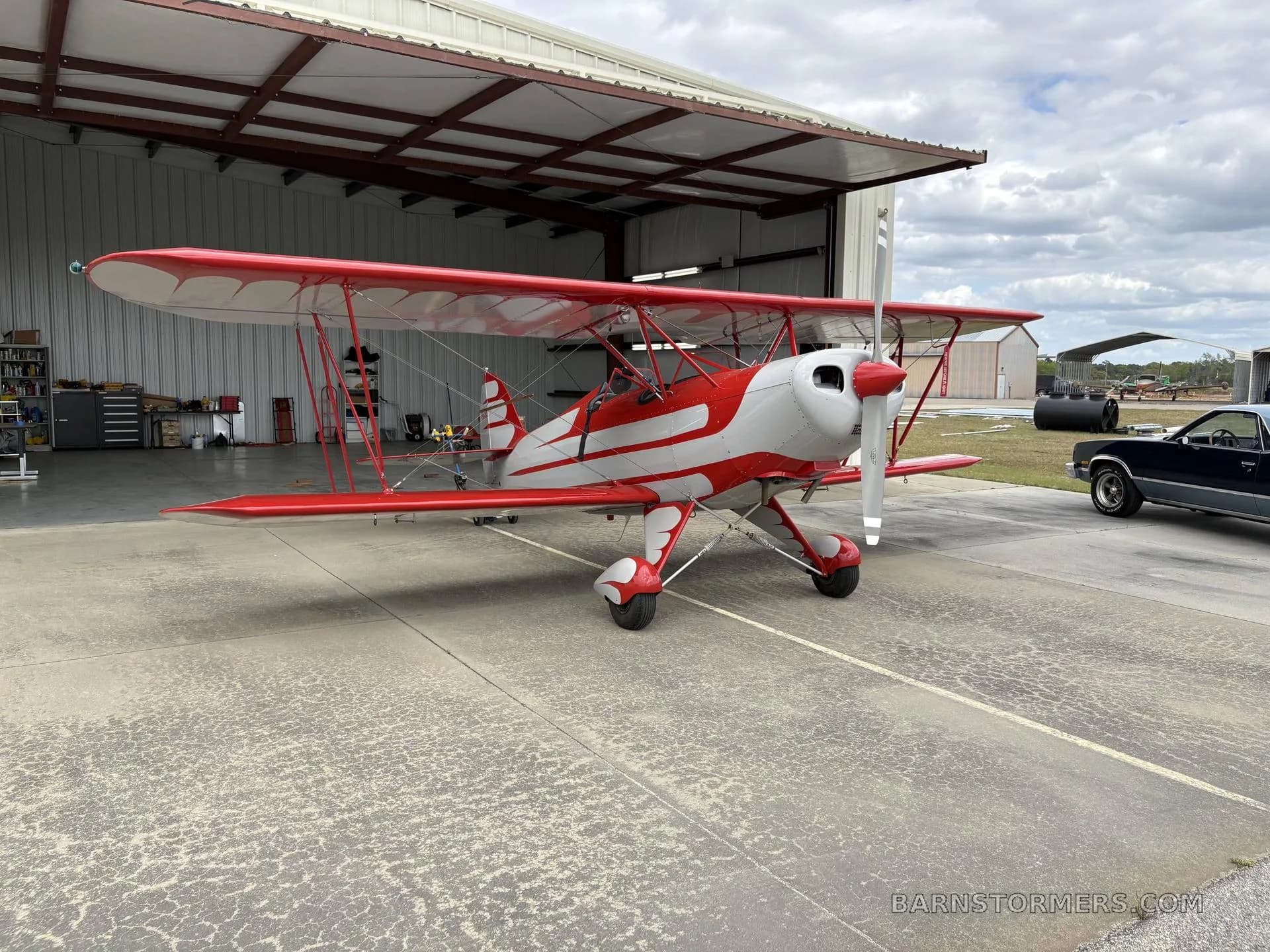 A red and white biplane with a radial engine is parked on concrete outside an open hangar.