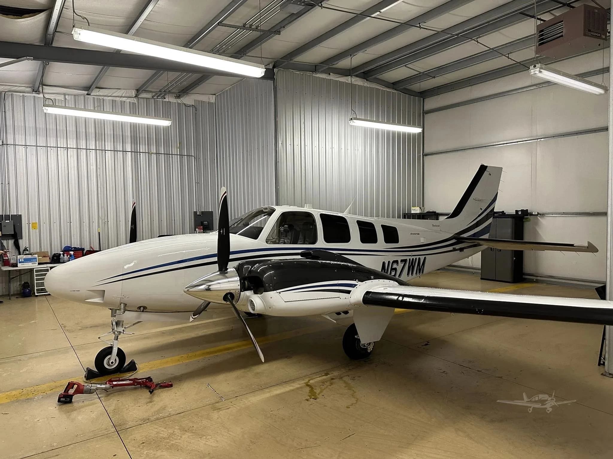 A white and blue Beechcraft Baron N67WM parked inside a hangar, viewed from the front-right.