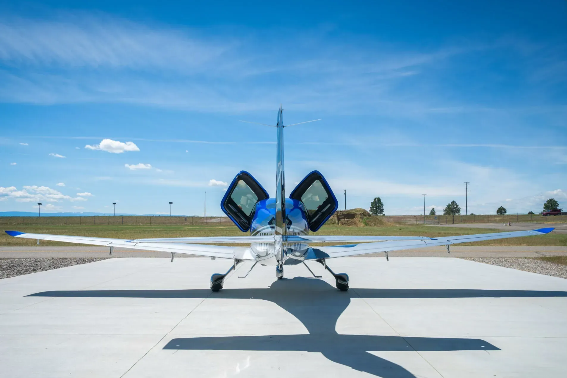 A rear view of the blue and silver Cirrus SR22T aircraft with its gull-wing doors open, parked on an airfield.