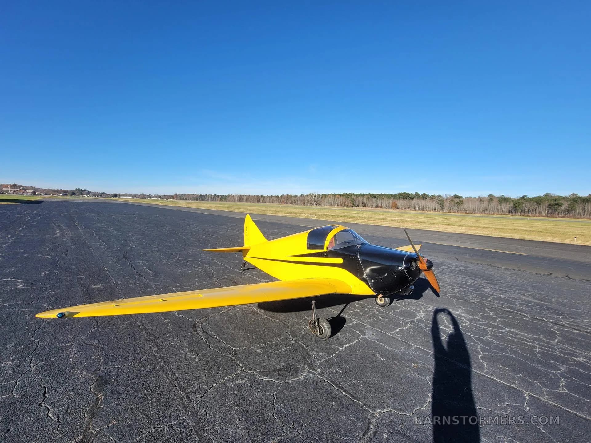 A yellow and black single-engine aircraft with a propeller, viewed from the front-right, on an asphalt surface under a clear blue sky.