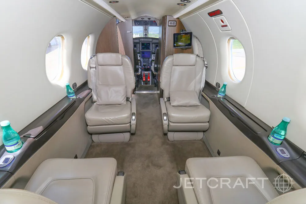 The interior cabin of a Pilatus PC-12 aircraft with light beige leather seats, looking forward towards the cockpit.