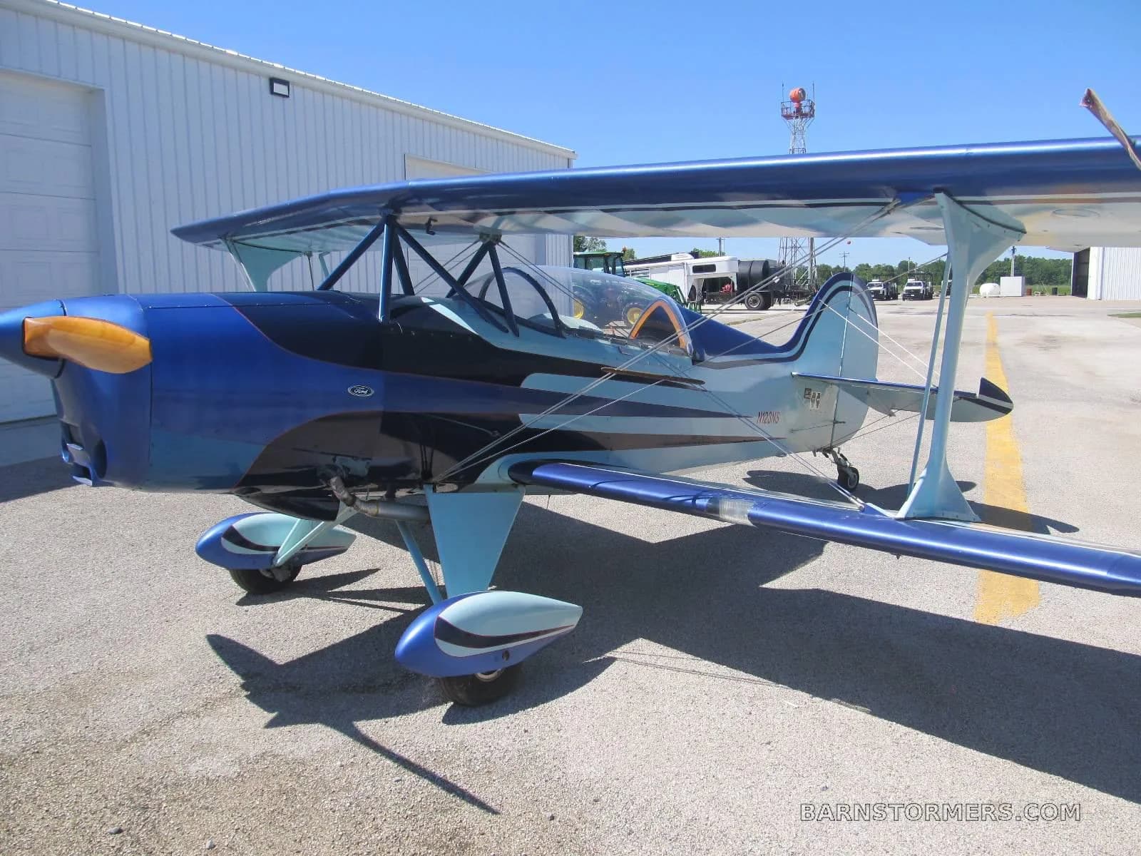 A blue and light blue biplane with an orange propeller spinner is parked on an asphalt apron next to a white building.