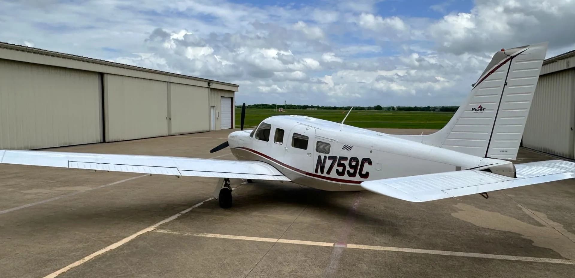 A white and tan Piper Saratoga II TC aircraft (N759C) parked on an asphalt ramp with hangars and a green field in the background.