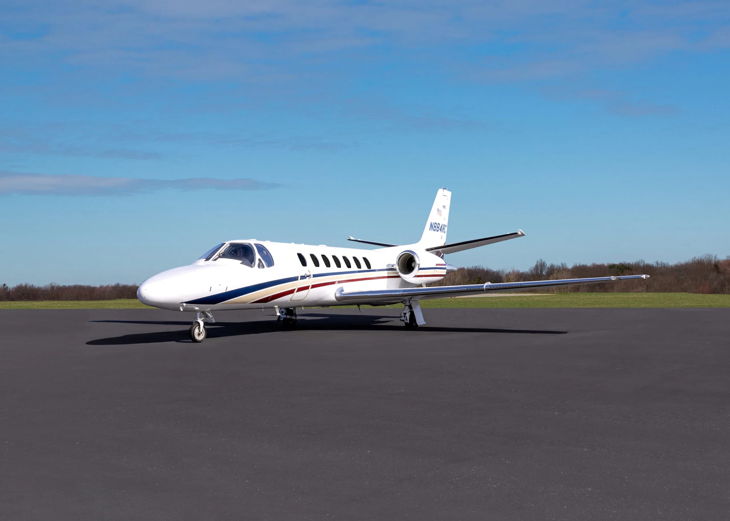 A white Cessna Citation business jet with blue, red, and gold stripes is parked on an asphalt tarmac under a clear blue sky.