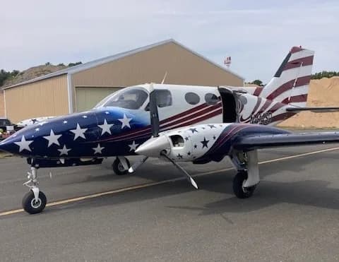A twin-engine propeller aircraft with a distinctive American flag paint scheme, featuring stars on the nose and stripes on the tail, parked on an airfield in front of a hangar.