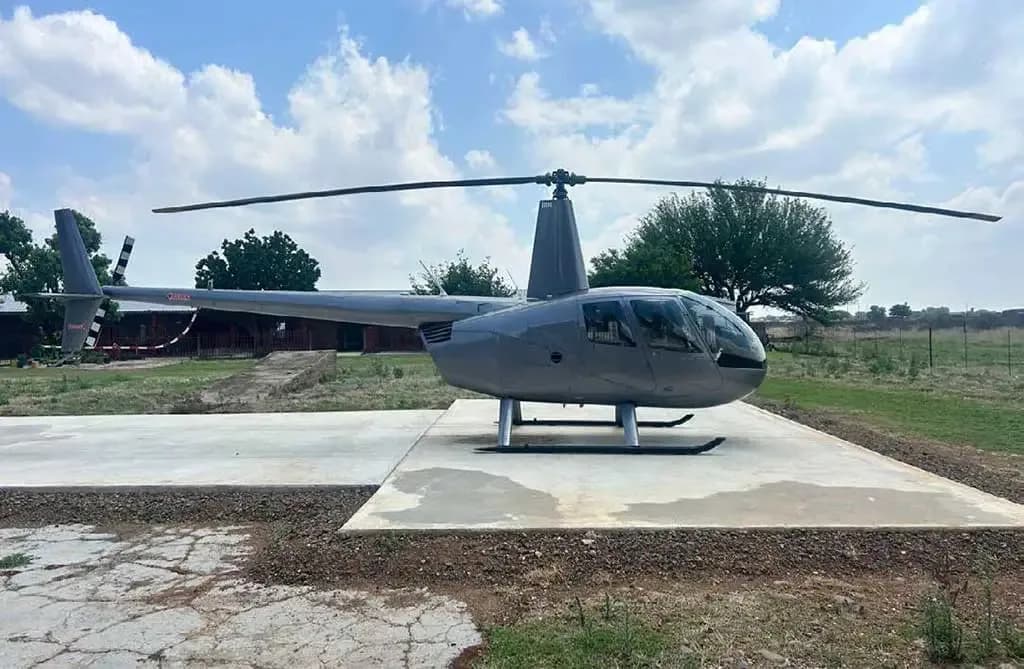 A side view of a silver Robinson R44 helicopter parked on a concrete pad in a rural setting.