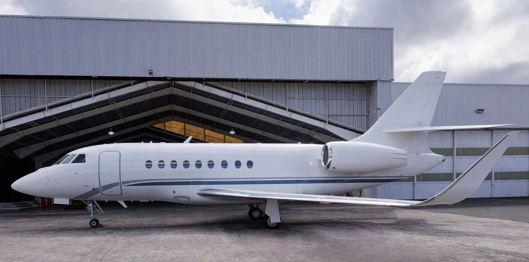 A white Dassault Falcon 2000LXS business jet with blue and grey stripes parked in front of a hangar.