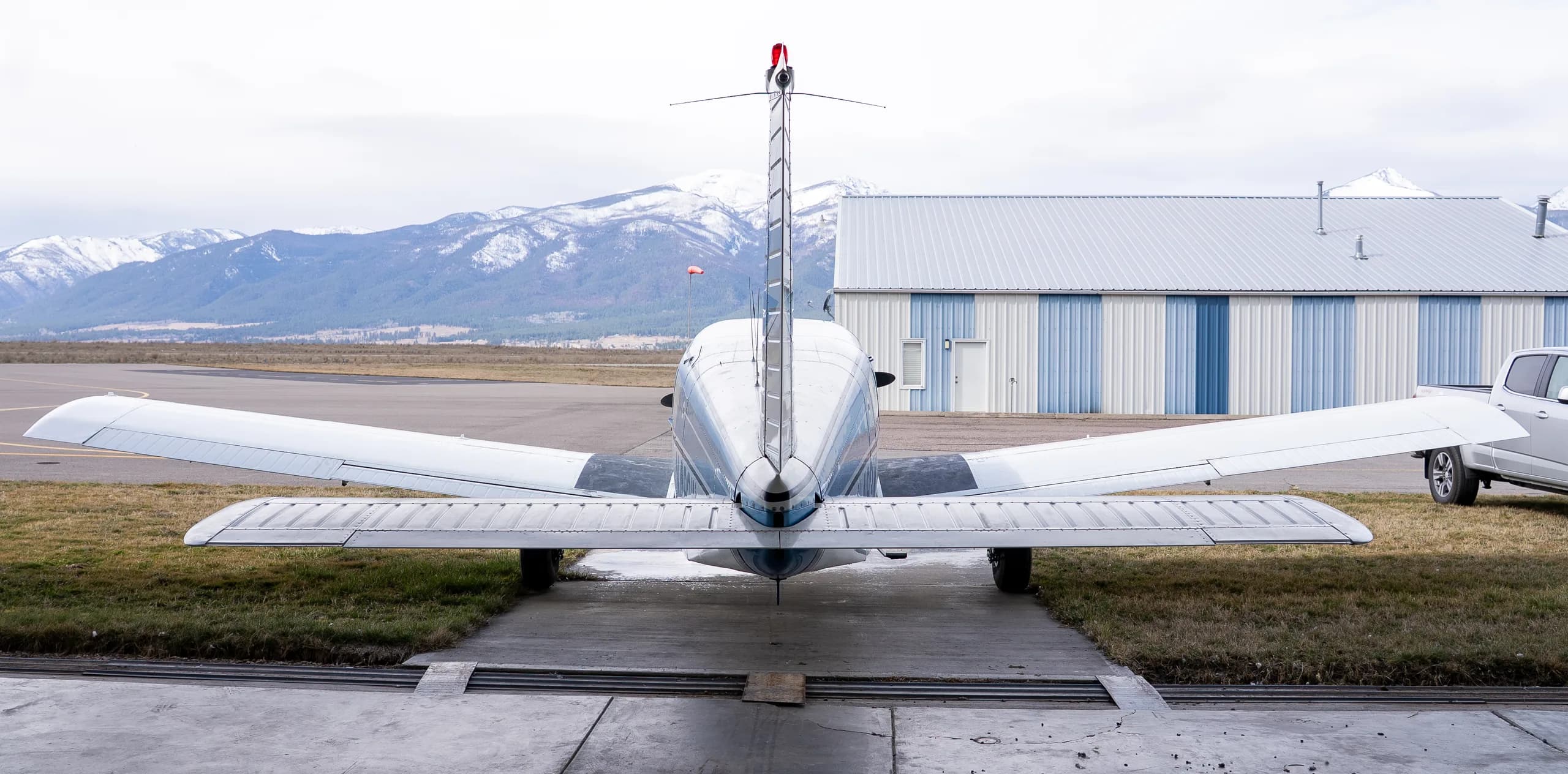 Rear view of a white and blue single-engine propeller aircraft N4193W parked outside a hangar with snowy mountains in the background.