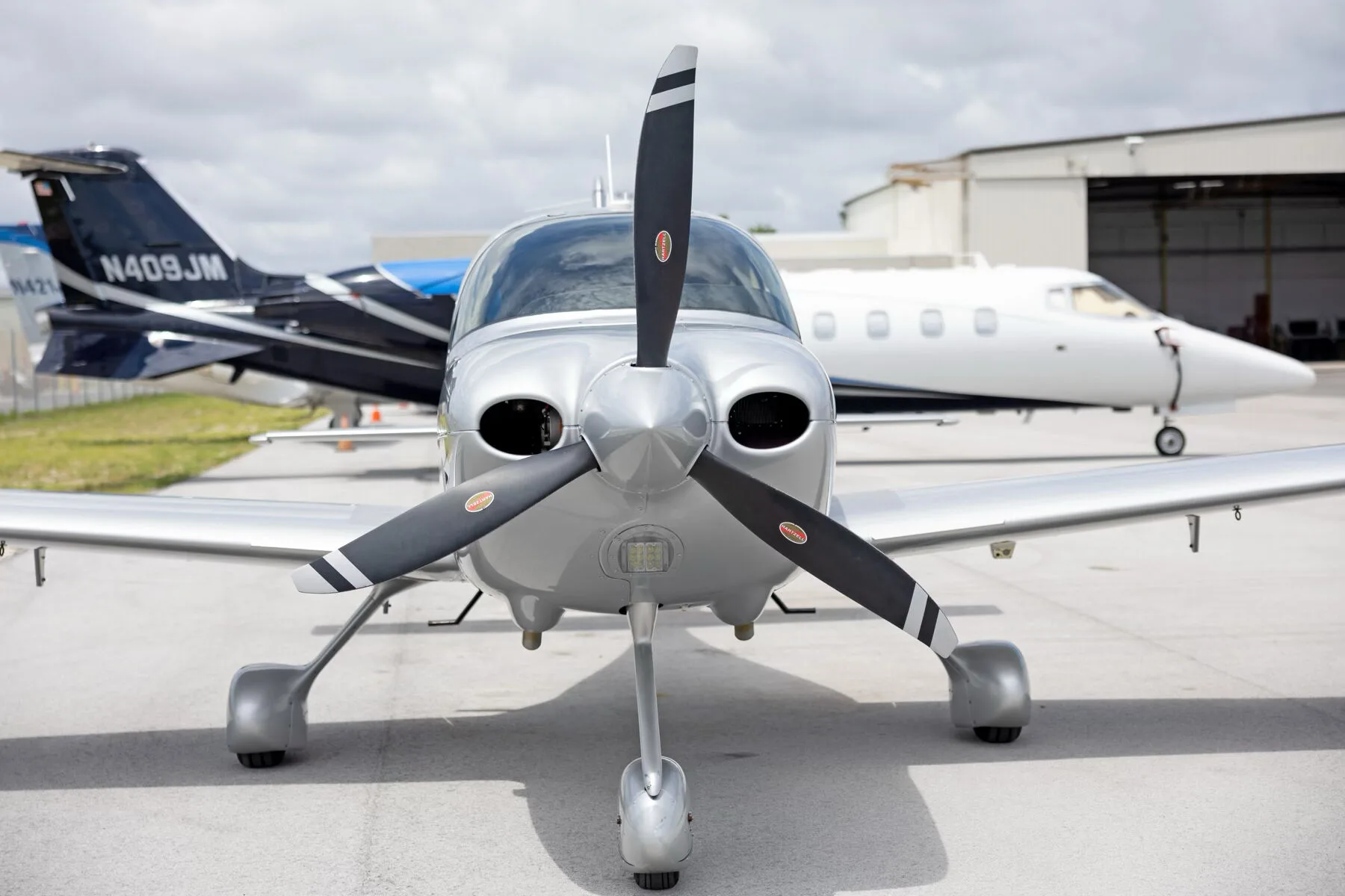 A head-on view of the silver Cirrus SR22 aircraft with its propeller, parked on an tarmac with other aircraft in the background.