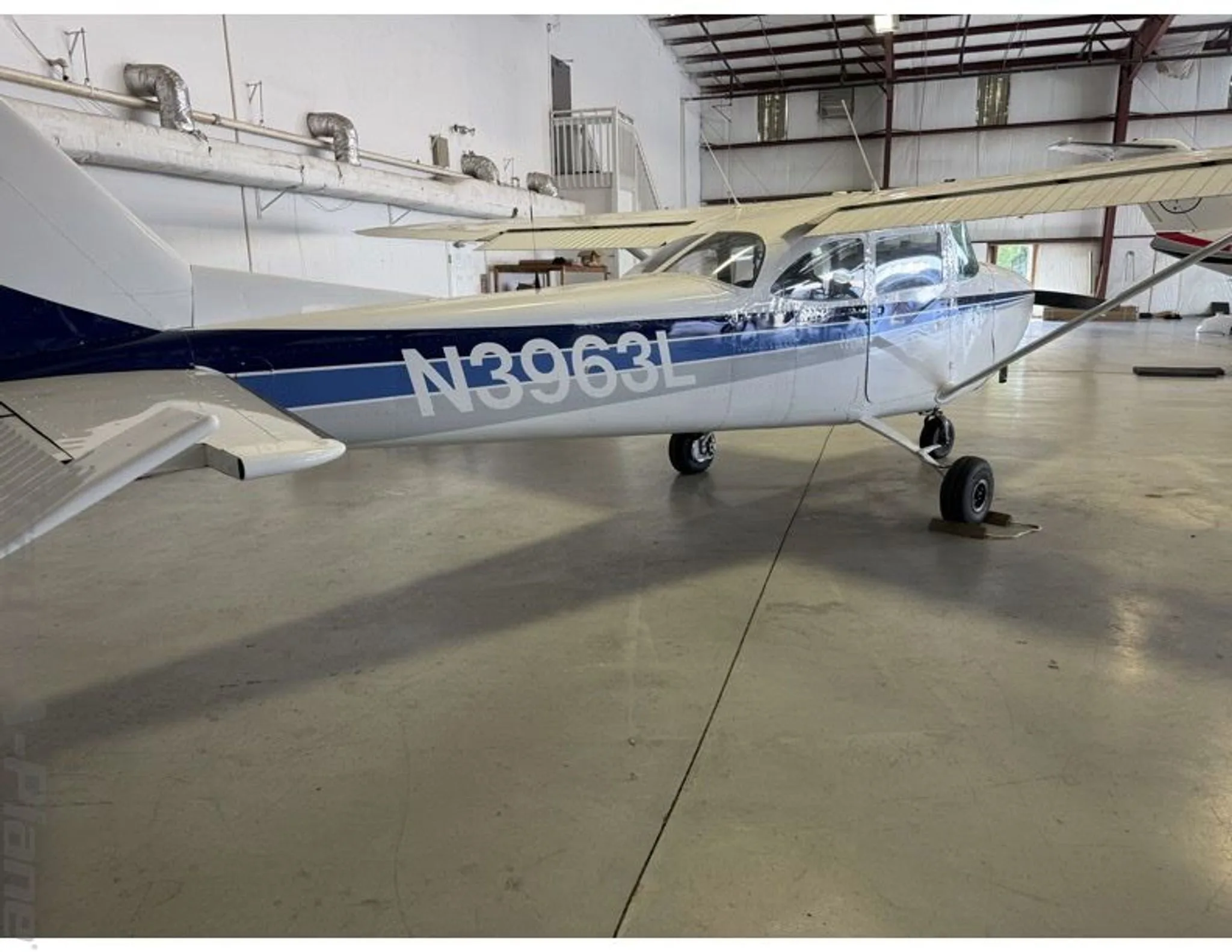 A side-rear view of a white Cessna 172 with blue and grey stripes, registration N3963L, parked in a hangar.