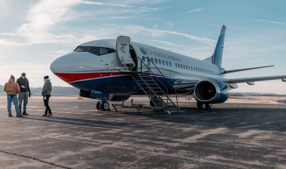 Side view of a Dynamic Aviation Boeing 737 aircraft on the tarmac with people nearby.