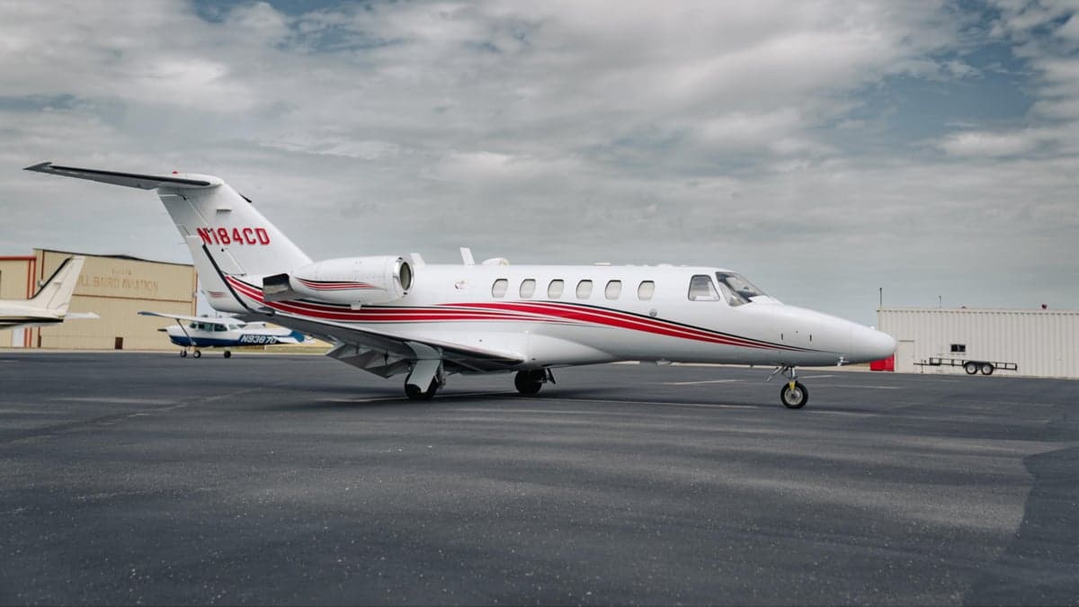 A white Citation CJ2 aircraft with red stripes is parked on the tarmac in front of a hangar.