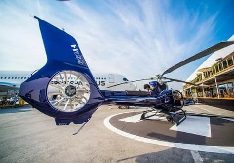 A blue H130 helicopter on a helipad with a large airplane in the background.