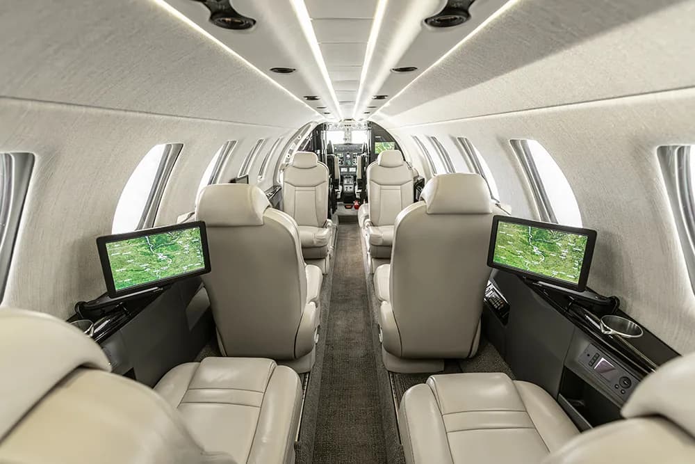 A wide view of the private jet's interior cabin, featuring multiple light grey seats and screens on the side panels.