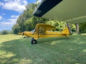 A yellow Cubcrafters aircraft is parked on the grass under the wing of another aircraft.