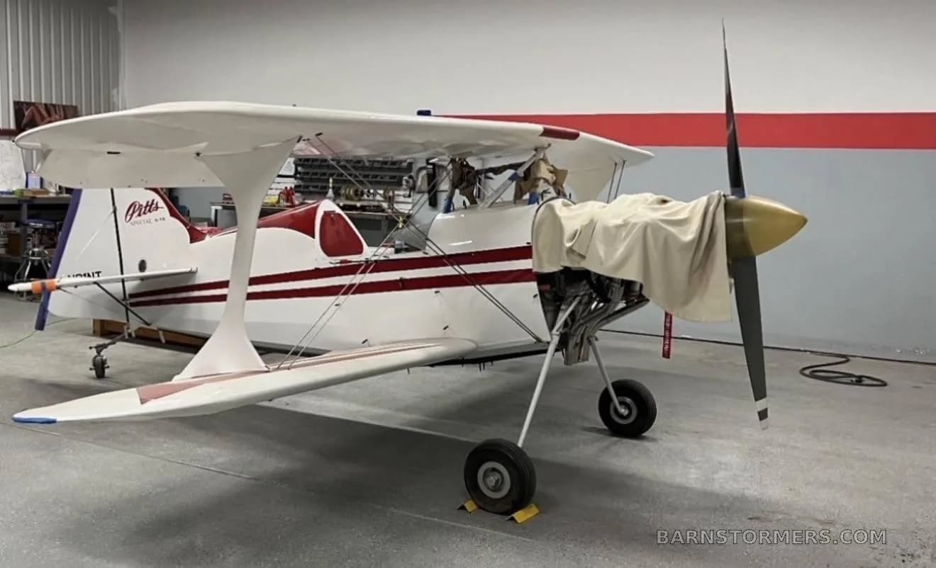 A white and red Pitts Special biplane with its engine exposed and propeller covered, parked in a hangar.