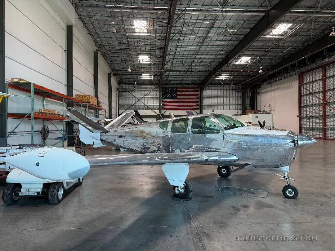 A bare metal, V-tail aircraft (N8558A) is parked in a hangar with an American flag in the background.