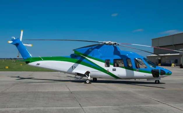 A blue, green, and white helicopter is parked on an airport apron with a hangar in the background.