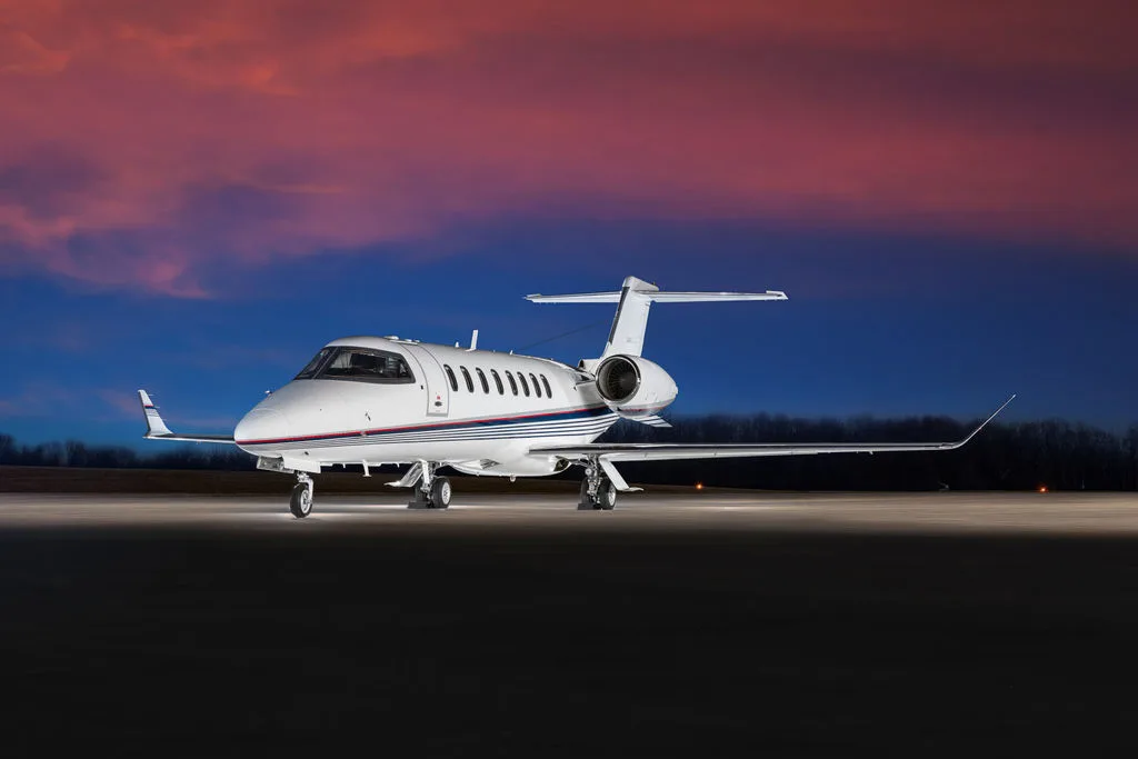 A white business jet with blue and red stripes on the fuselage is parked on a tarmac at sunset.