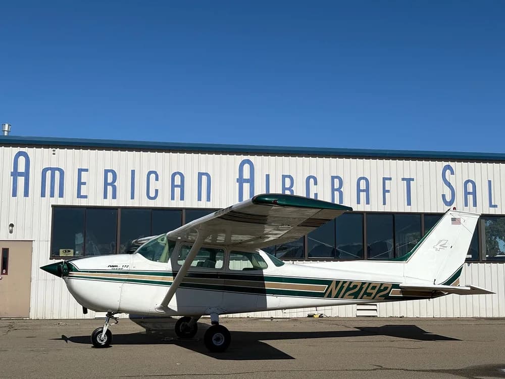 A white Cessna 172 with green and gold stripes parked on an airfield in front of a hangar, viewed from the left side.