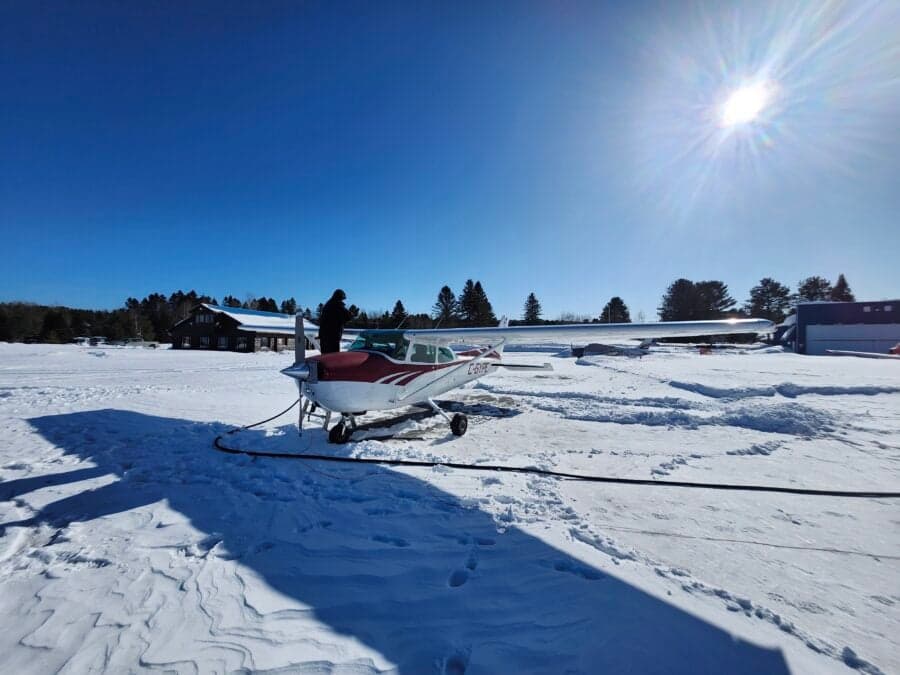 A person stands near the propeller of a white and red Cessna 172 (C-GYPE) parked in snow on a sunny day.