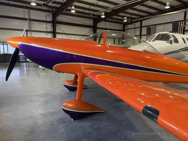 A close-up front view of the orange and purple single-engine aerobatic aircraft's nose and wing in a hangar.