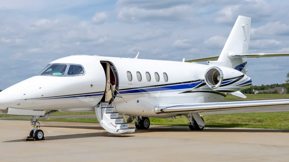 A white private jet with blue and black stripes is parked on the tarmac with its main door open and stairs extended.