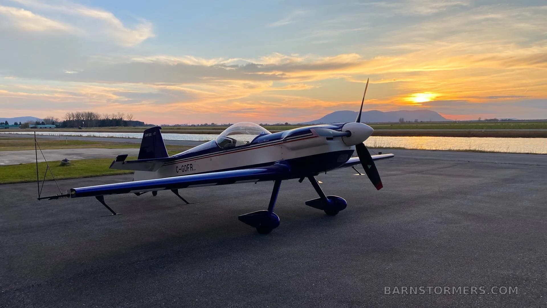 A blue and white CAP 10B aerobatic aircraft is parked on an airfield at sunset, with mountains and water in the background.