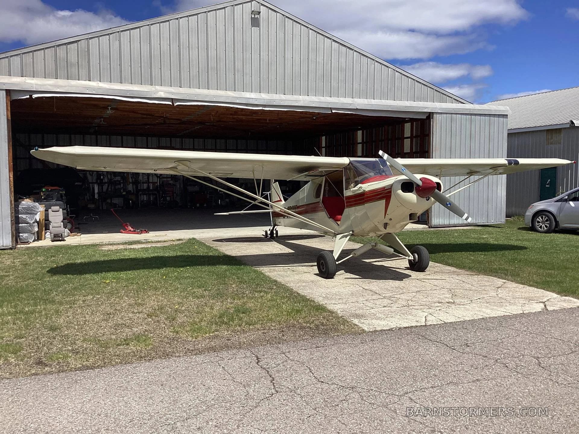 A cream and red high-wing aircraft with a propeller parked on a concrete pad in front of an open hangar on a sunny day.
