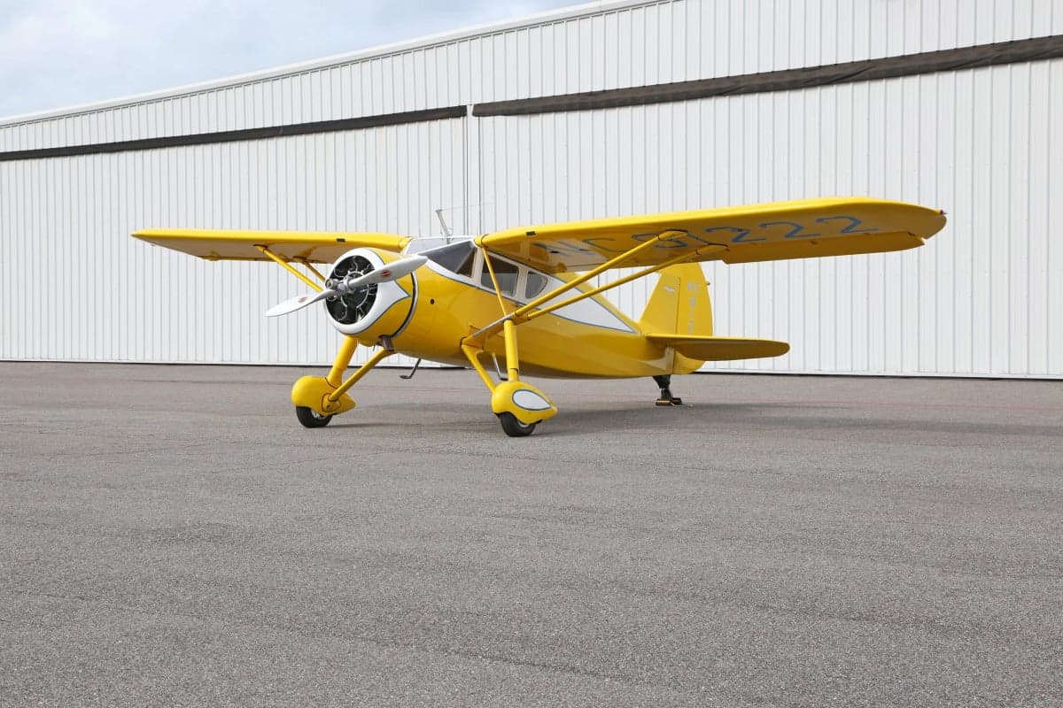 A yellow and white Stinson Reliant parked on the tarmac in front of a hangar.