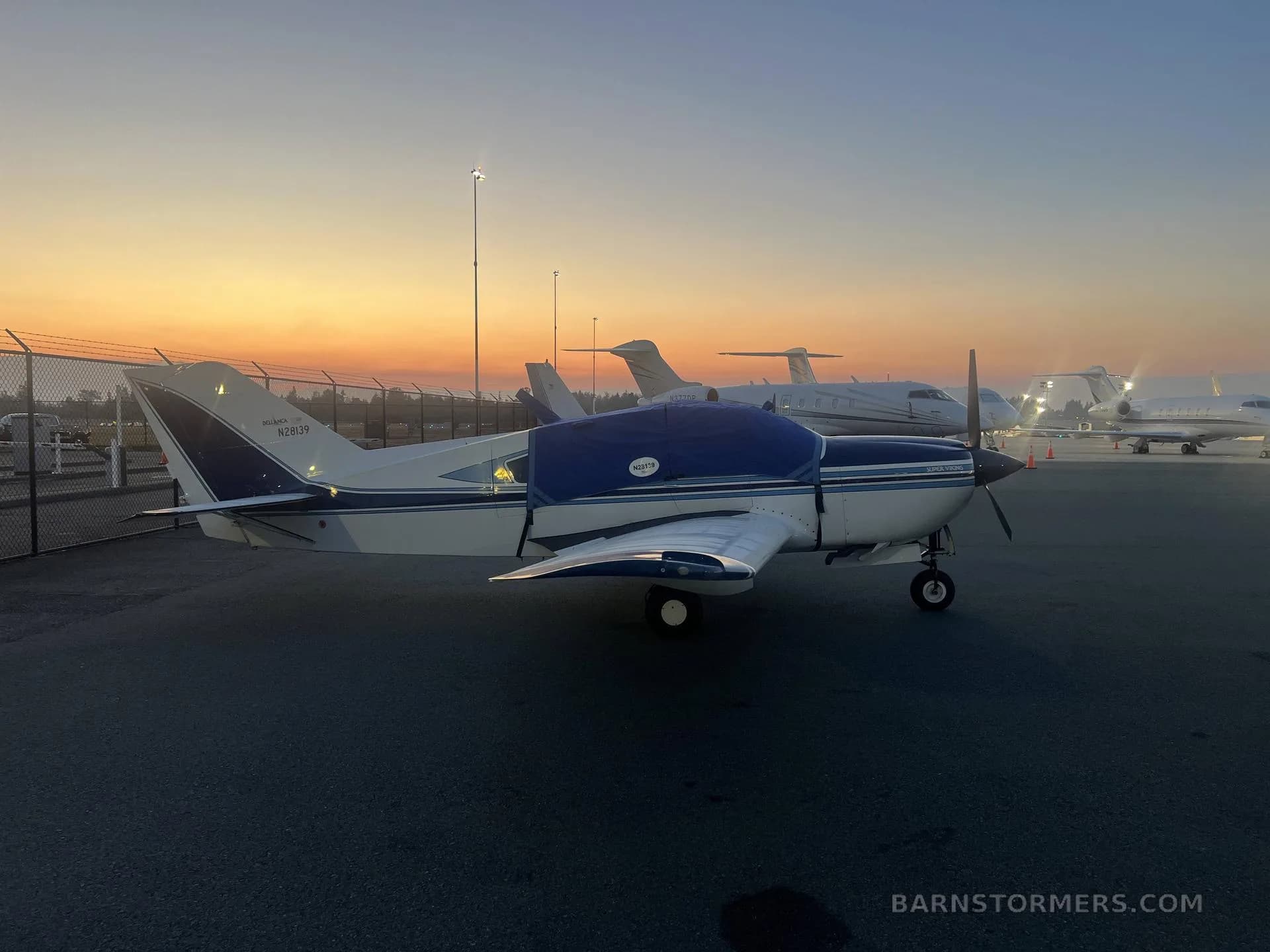 A white and blue Bellanca Viking aircraft with a blue engine cover parked on an airport tarmac at sunset.