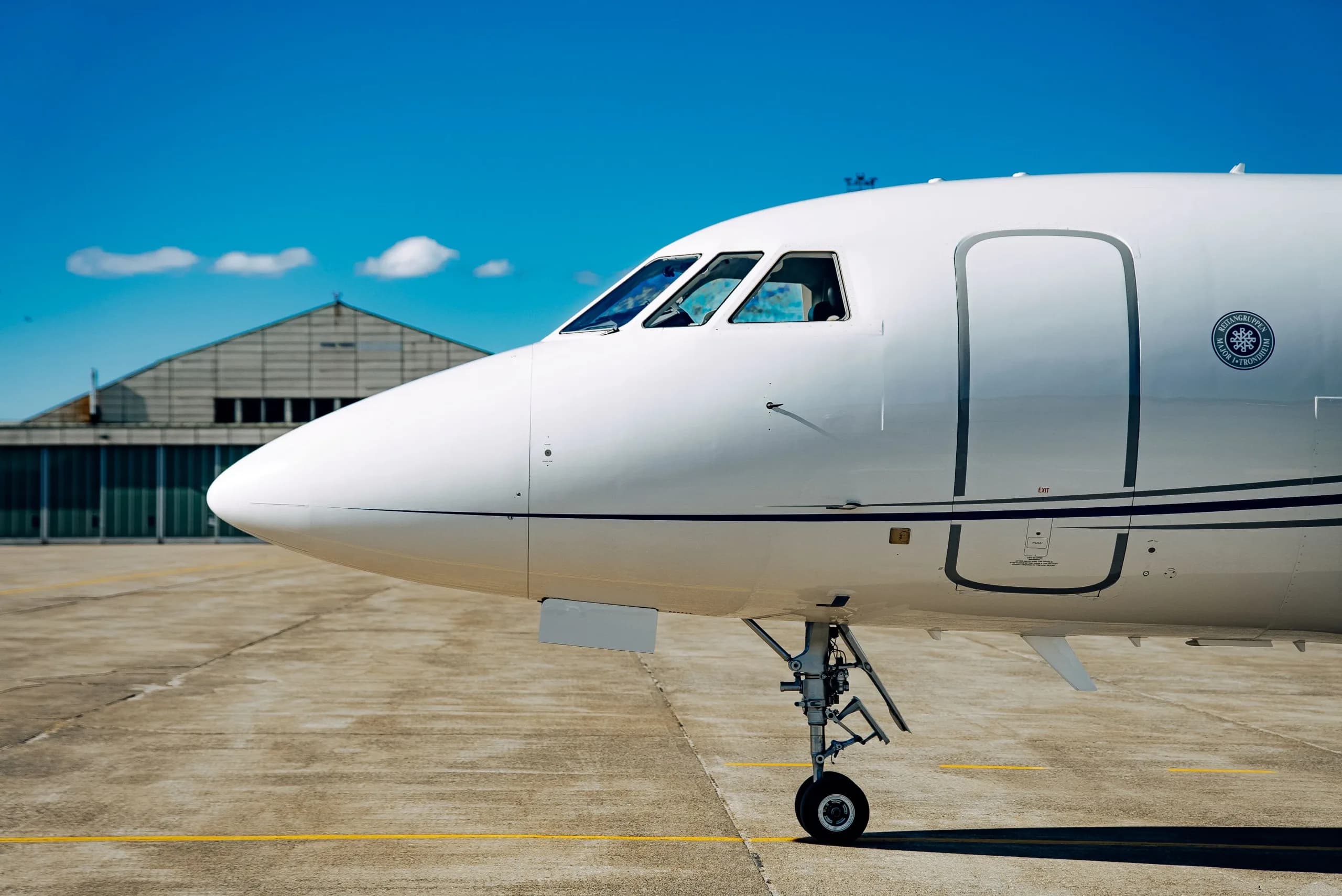 A close-up view of the nose and cockpit of a white business jet with a hangar in the background.