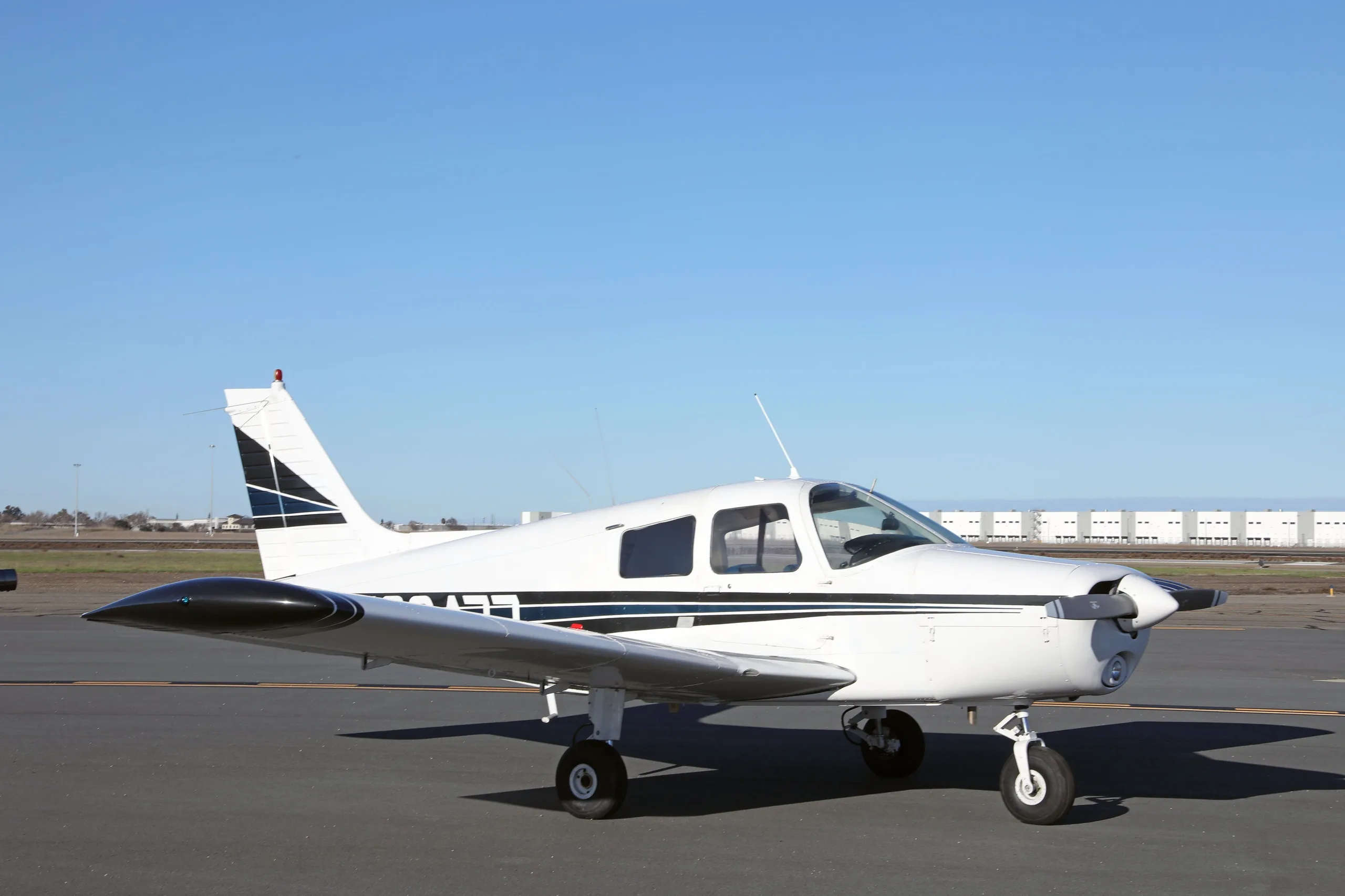 The white Piper Cherokee N38477 with blue and black stripes is seen from the left side on an airfield.
